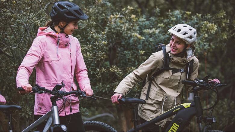 Två personer med cyklar står utomhus och pratar glatt med varandra. De bär hjälmar och regnjackor, e